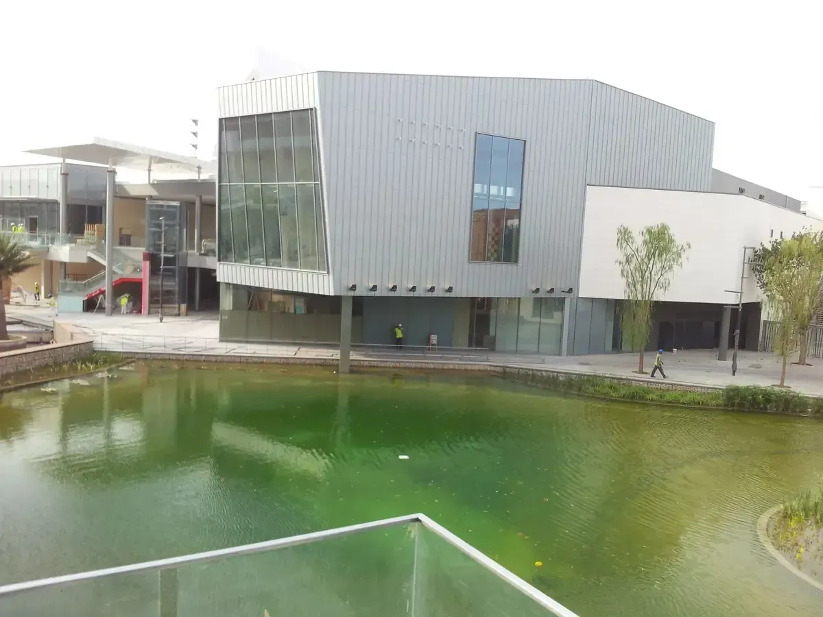 Edificio moderno con fachada metálica y grandes ventanales, reflejados en un estanque verde.