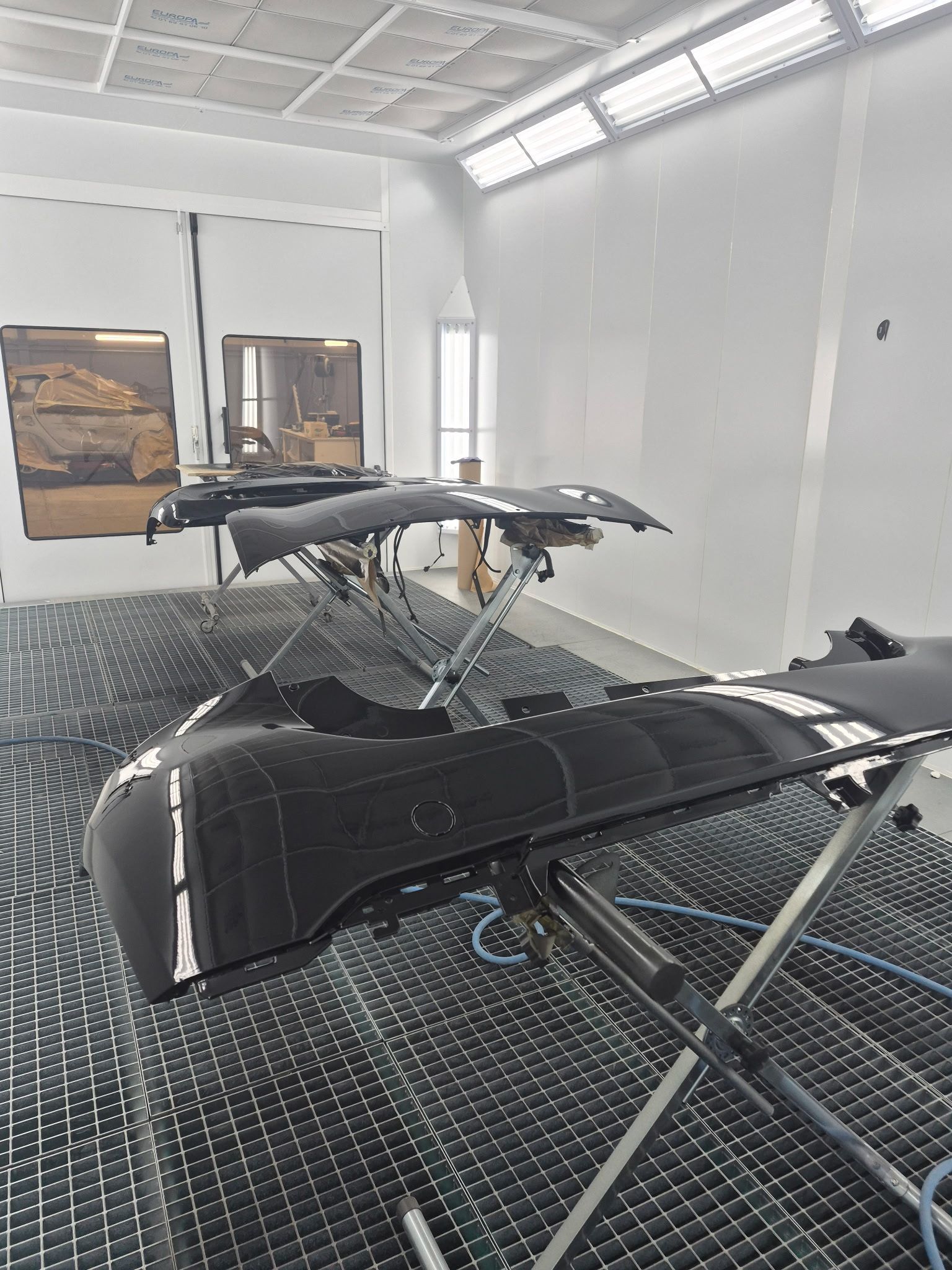 Deux pare-chocs de voiture noirs montés sur des supports métalliques à l'intérieur d'une cabine de peinture automobile blanche et éclairée.