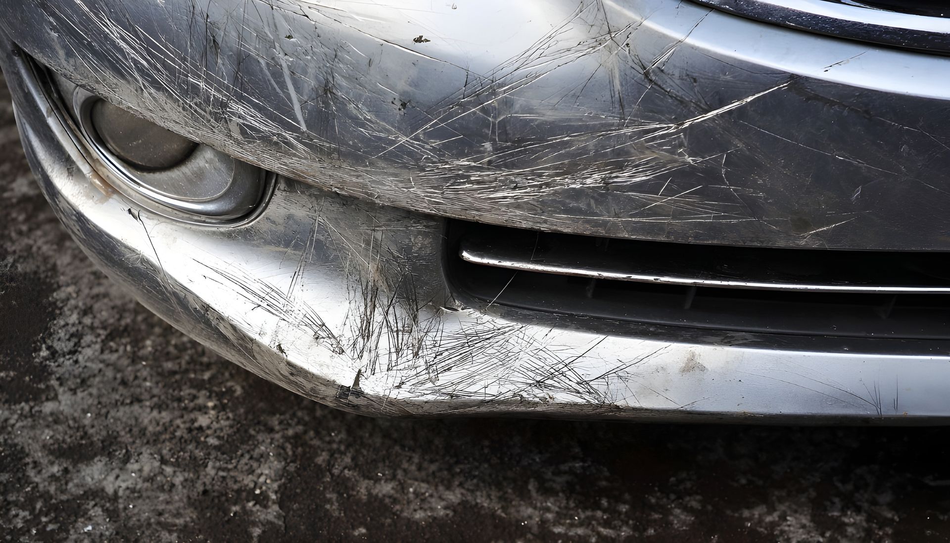 Gros plan sur un pare-chocs de voiture argenté présentant de profondes éraflures blanches, des rayures marquées et des marques d'usure foncées.