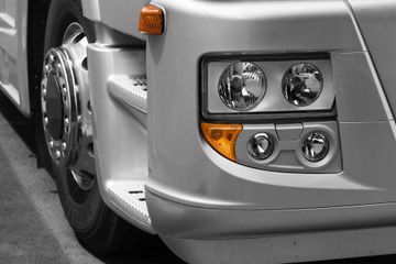 Gros plan sur la roue avant, les marchepieds et le bloc optique avant d'un camion argenté, avec ses doubles phares circulaires et ses clignotants ambrés.