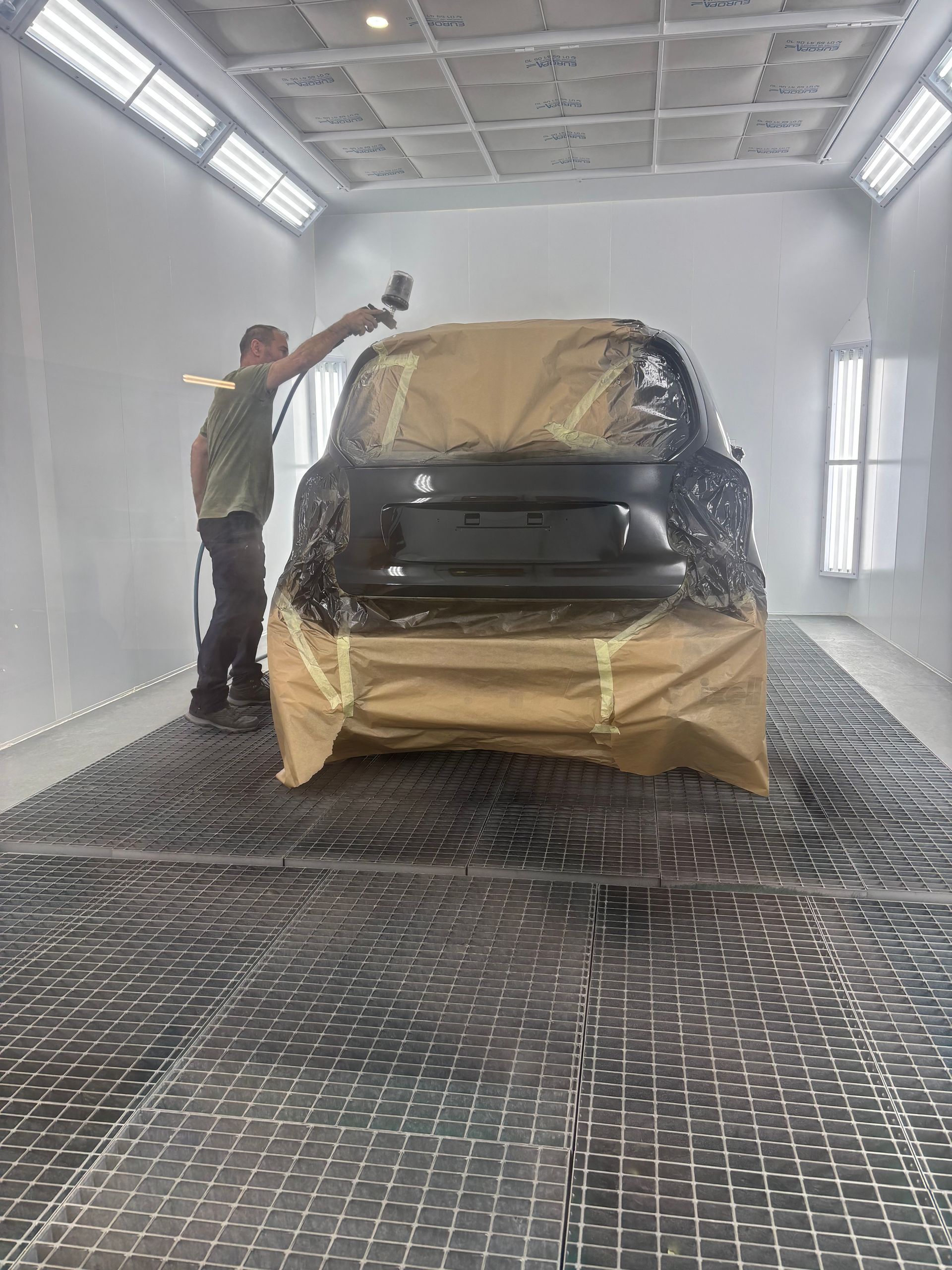 Une personne dans une cabine de peinture en train de peindre l'arrière d'une voiture recouverte d'un papier protecteur.