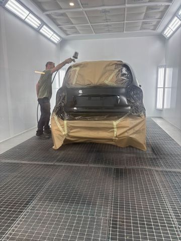 Une personne utilise un pistolet à peinture pour appliquer de la peinture à l'arrière d'une voiture à l'intérieur d'une cabine de peinture automobile bien éclairée.