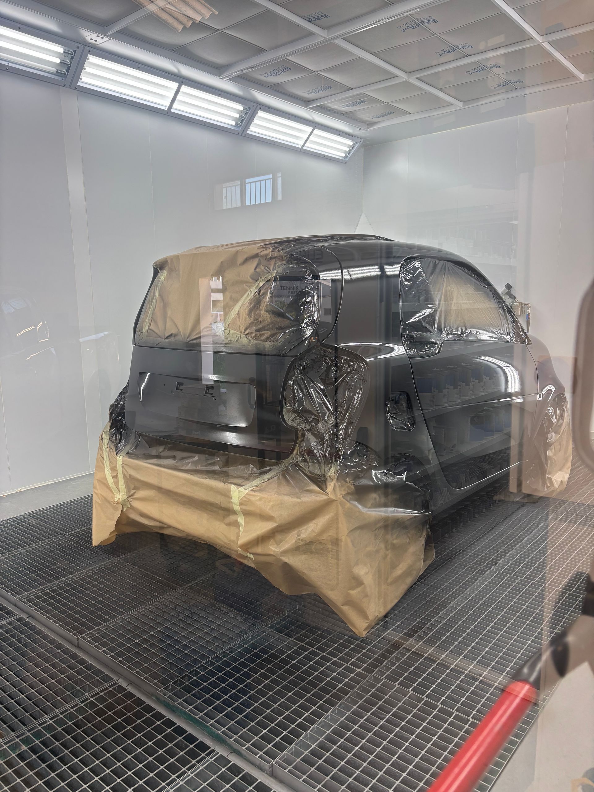 Une voiture sombre est garée à l'intérieur d'une cabine de peinture blanche éclatante d'un atelier de carrosserie, les vitres et le pare-chocs étant masqués par du ruban adhésif beige.