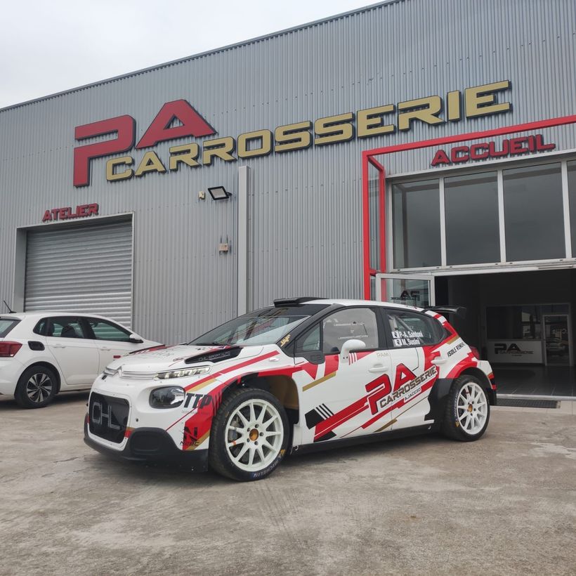 Une voiture de rallye blanche et rouge garée devant l'entrée de l'atelier de la carrosserie PA.