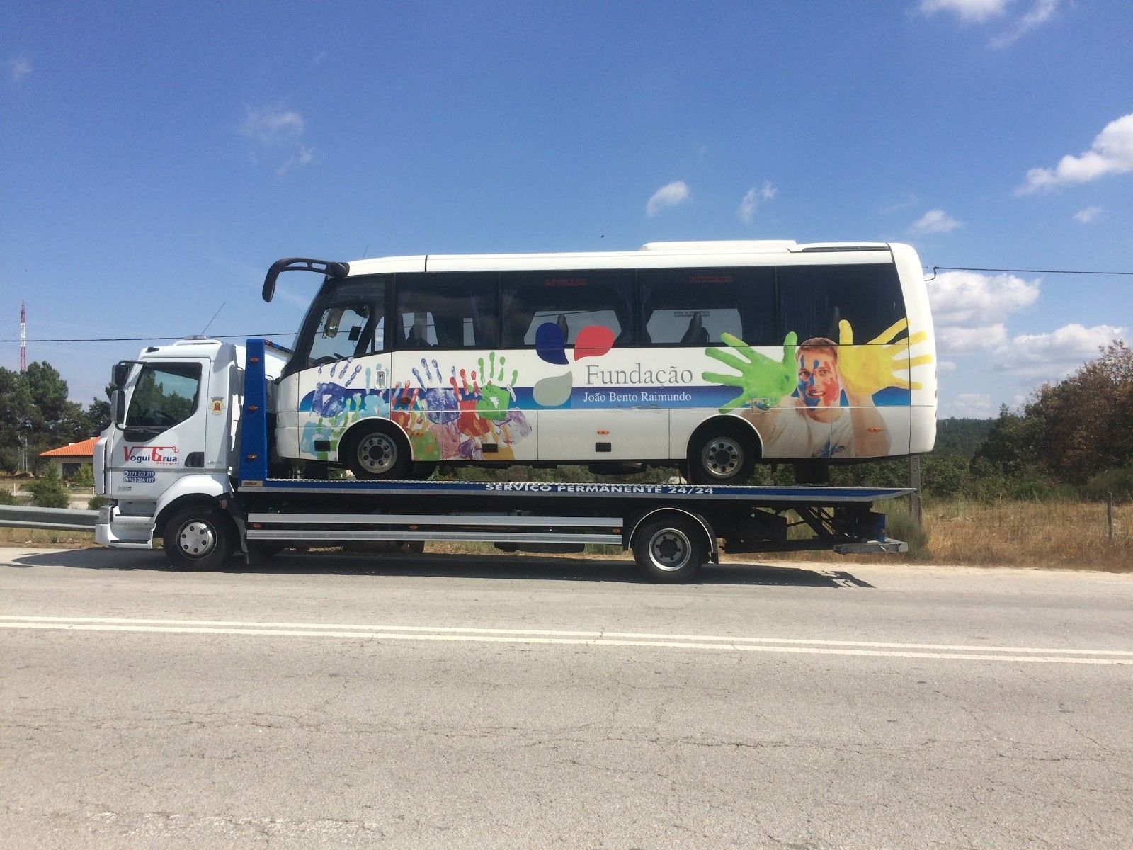 Caminhão guincho transportando um ônibus branco e colorido com decorações de mãos e desenhos animados em um dia ensolarado.