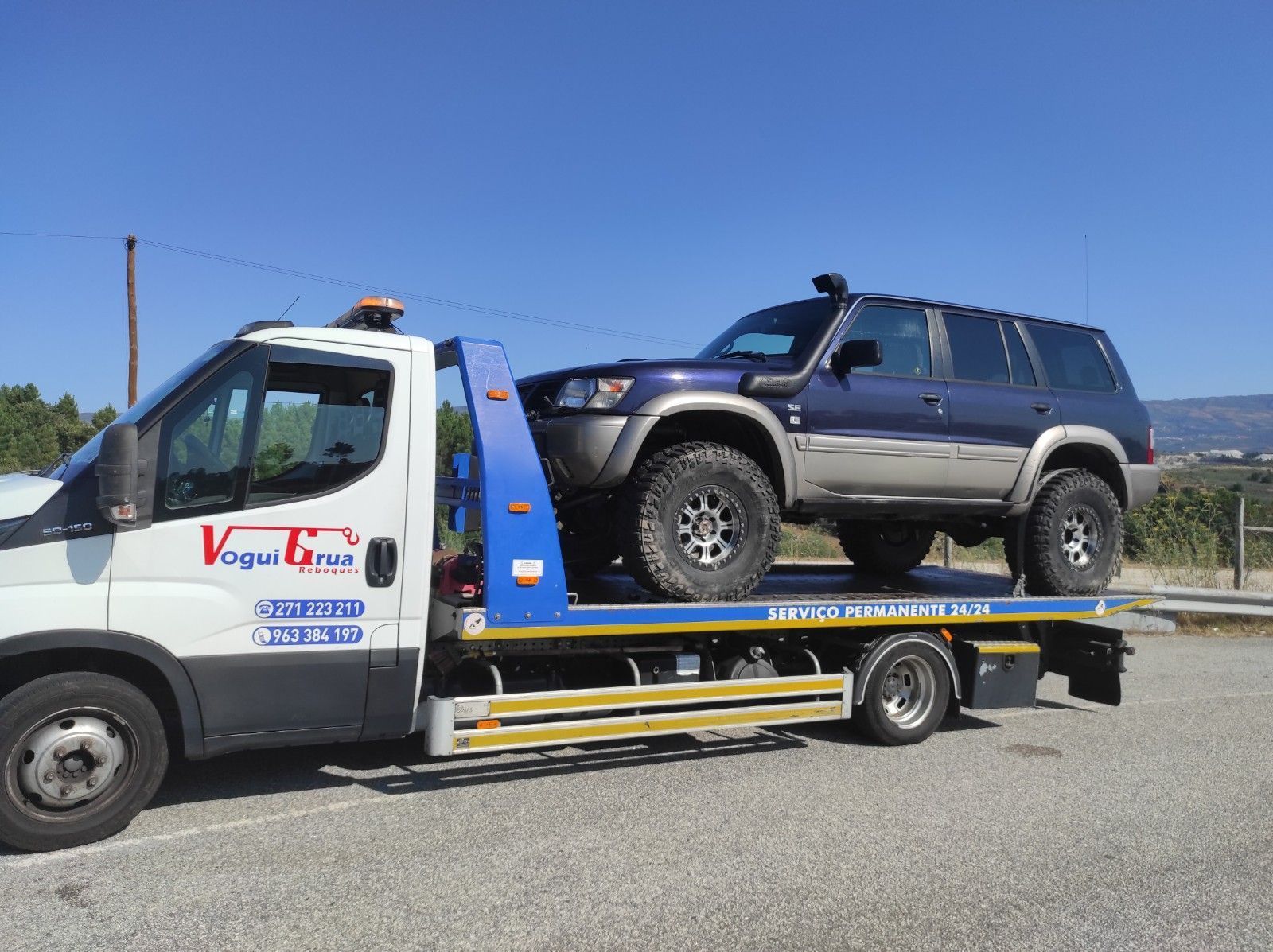 Um SUV azul sendo rebocado por um caminhão-guincho branco em um dia ensolarado.