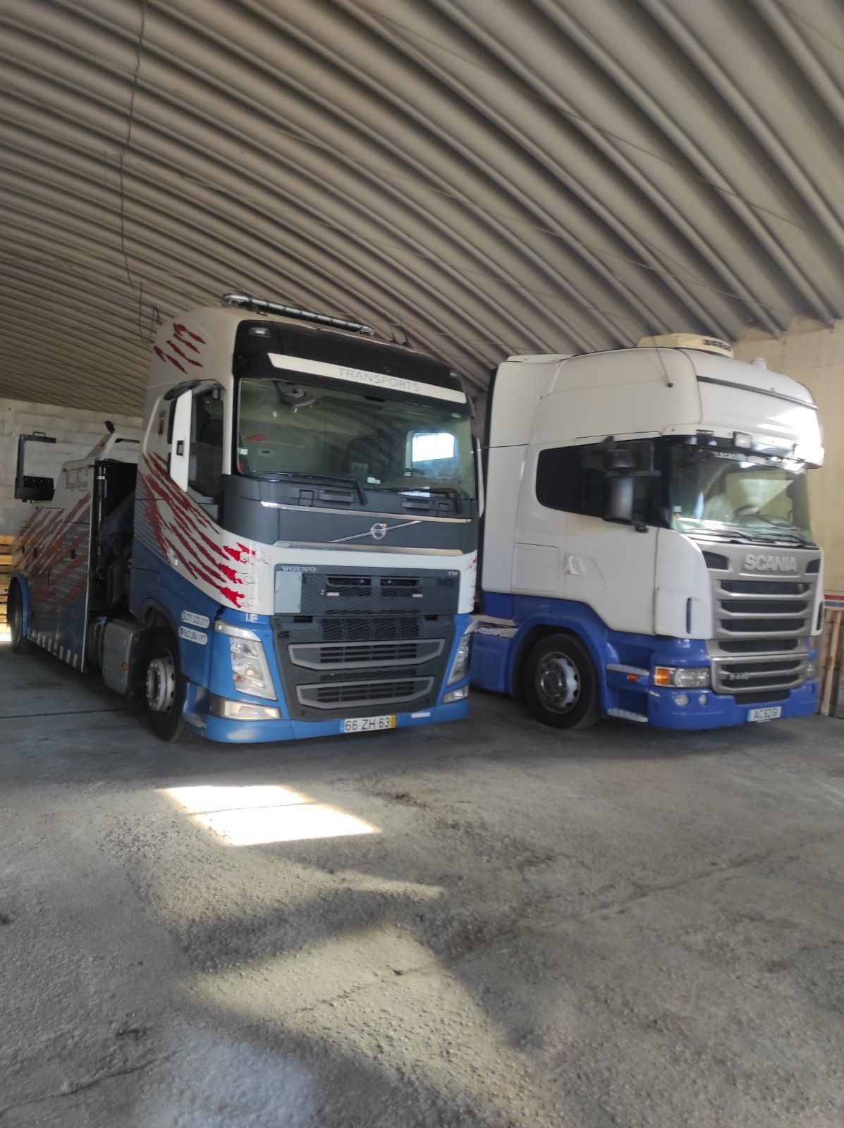 Dois caminhões semirreboque estacionados dentro de um galpão industrial leve. Um é azul e branco, o outro é branco e azul.