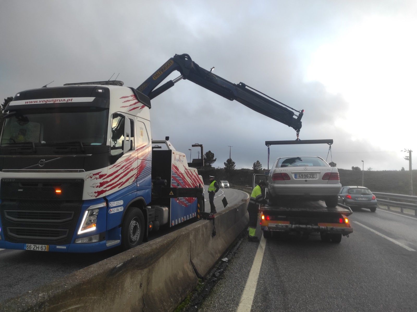 Um guincho está içando um carro para dentro de sua carreta em uma rodovia, sob um céu nublado.