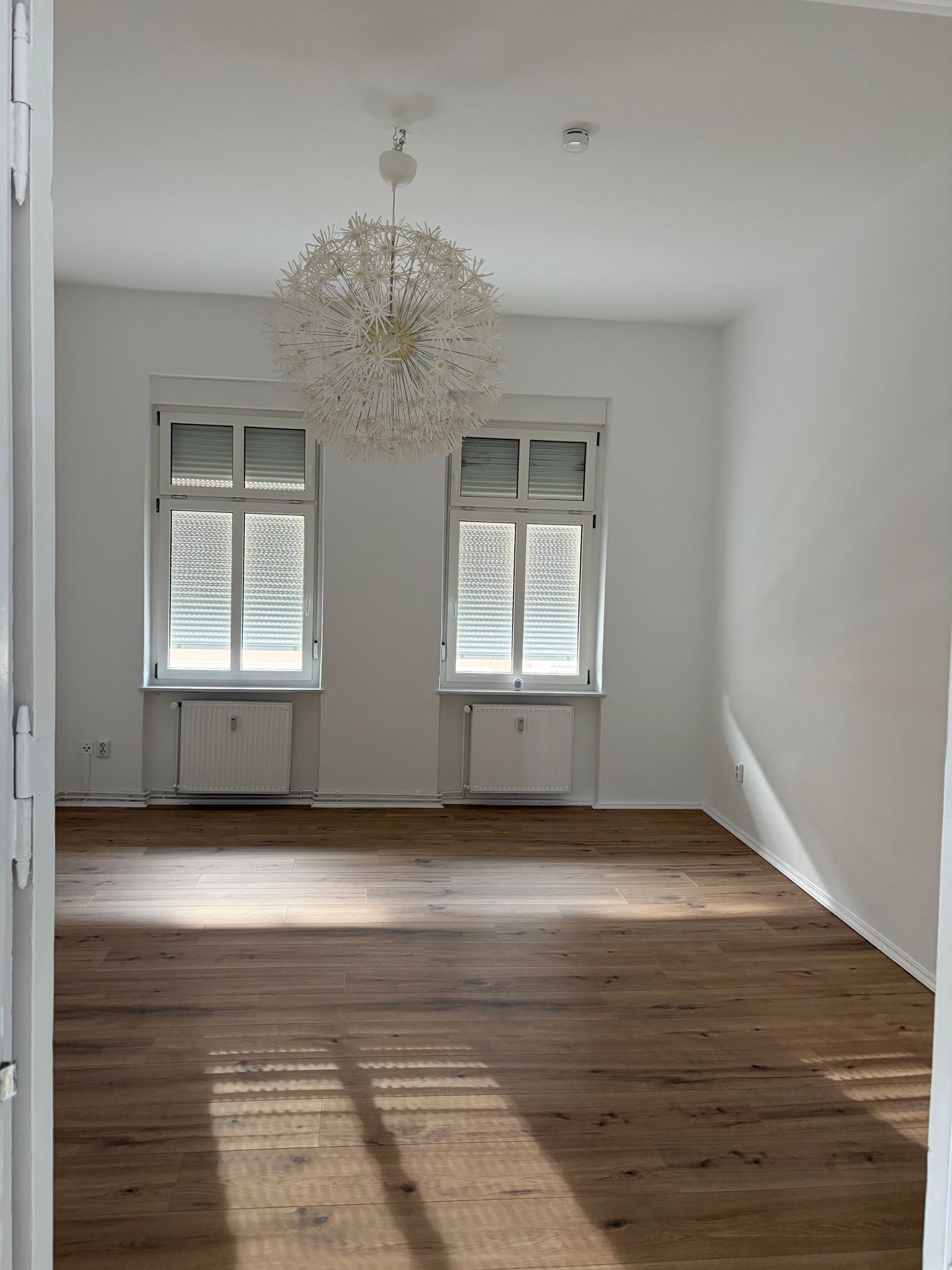 Leerer weißer Raum mit Holzboden, zwei Fensterläden und einem Kronleuchter.