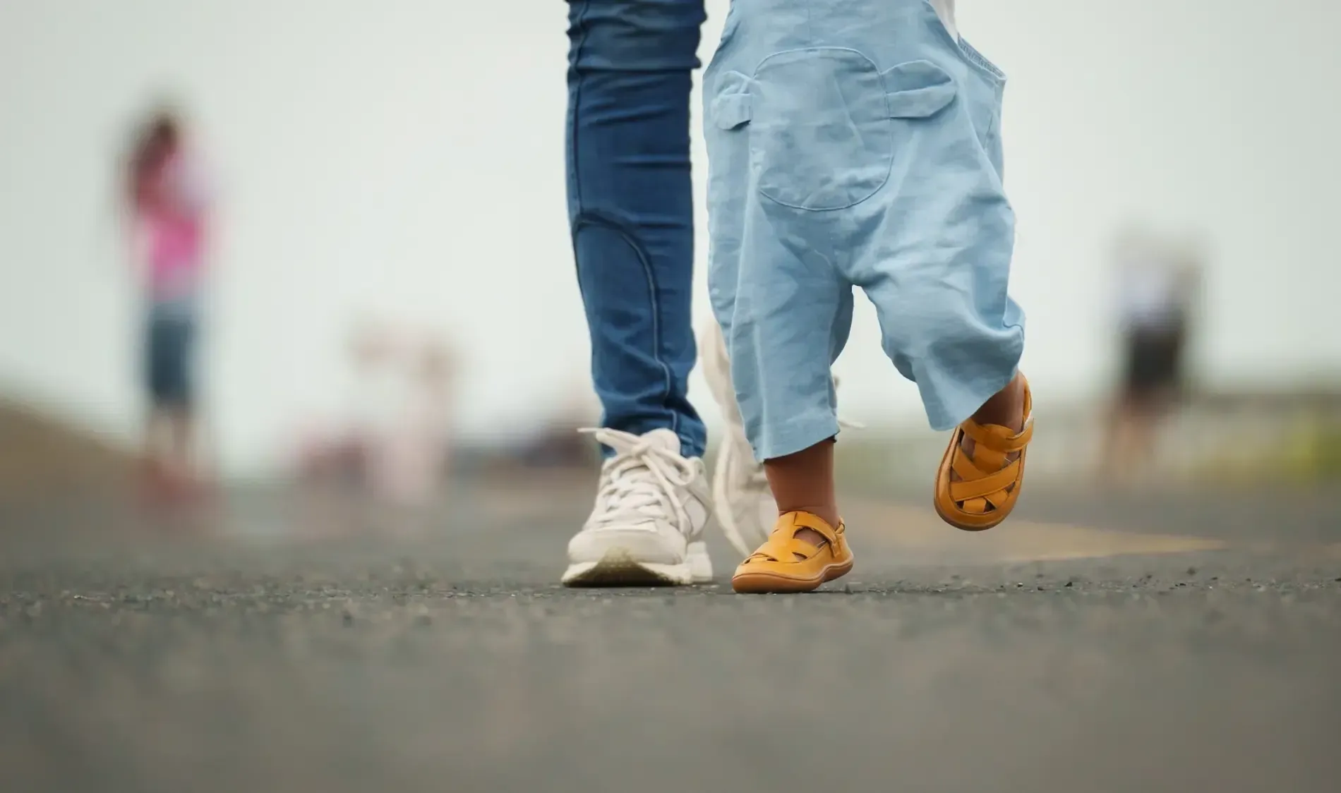 Pies de un niño con sandalias marrones caminando junto a un adulto con zapatillas blancas sobre una superficie pavimentada.