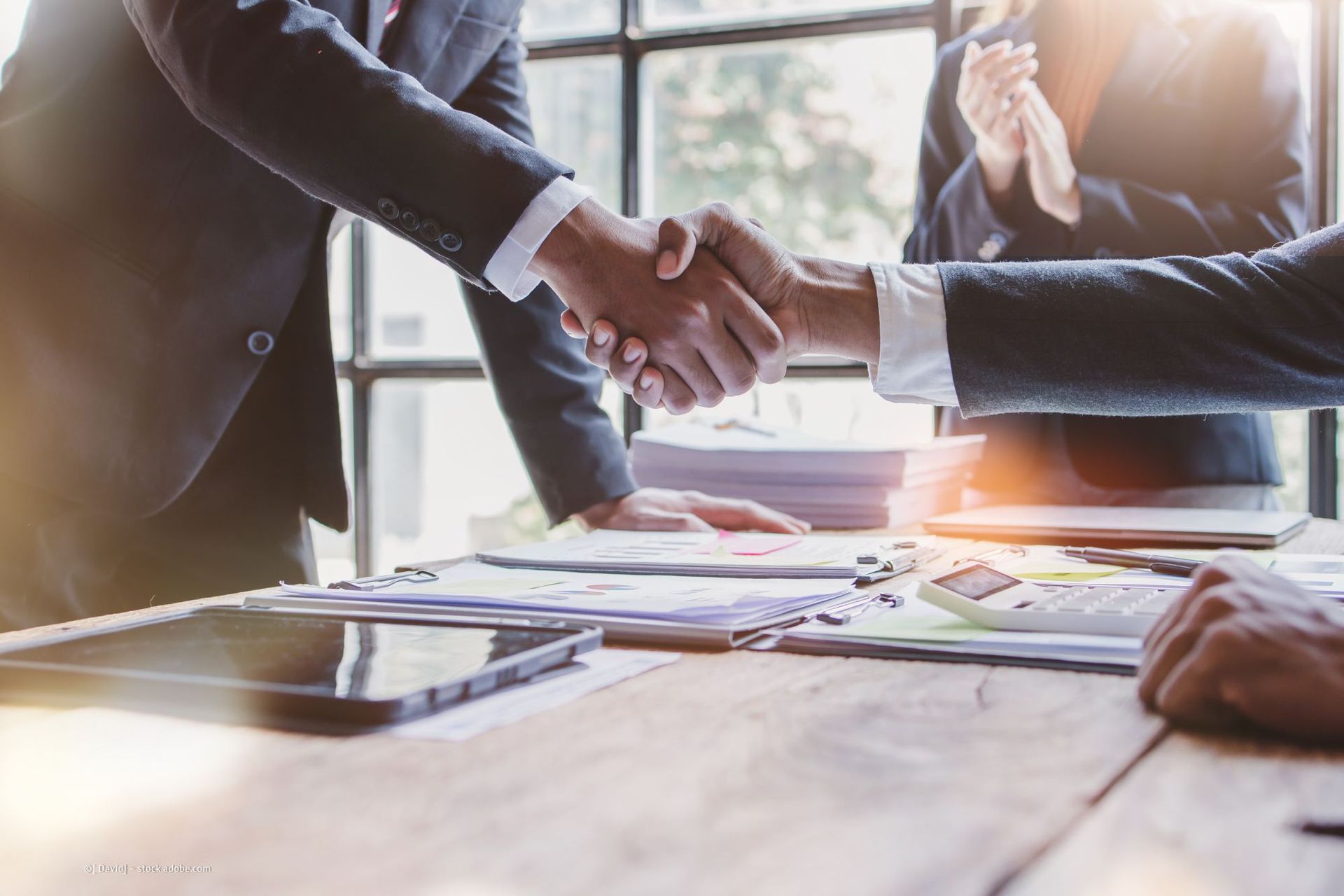 Zwei Personen in Anzügen geben sich die Hand, eine Person im Hintergrund klatscht