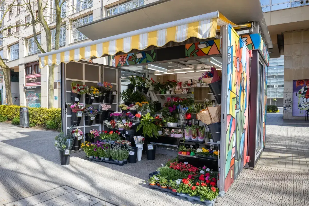 Quiosco de floristería con coloridos ramos y plantas en macetas bajo un toldo amarillo en una acera soleada.
