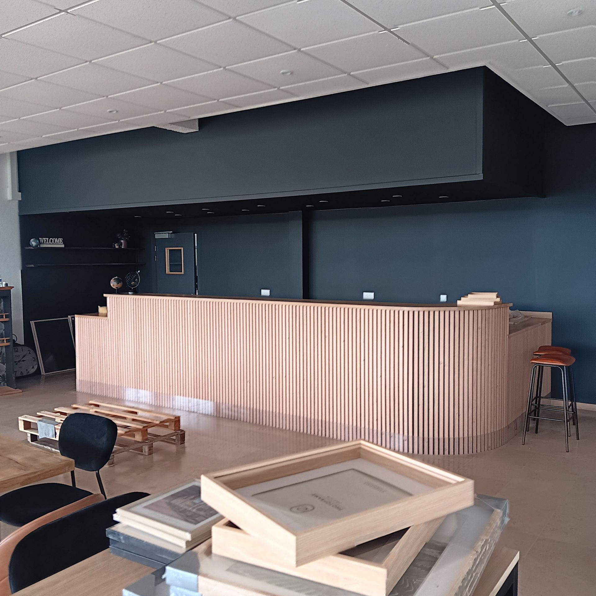 Une personne en tenue de travail et casque de chantier bleu utilise un rouleau à peinture pour peindre un mur en blanc dans une pièce vide, à l'aide d'une échelle.