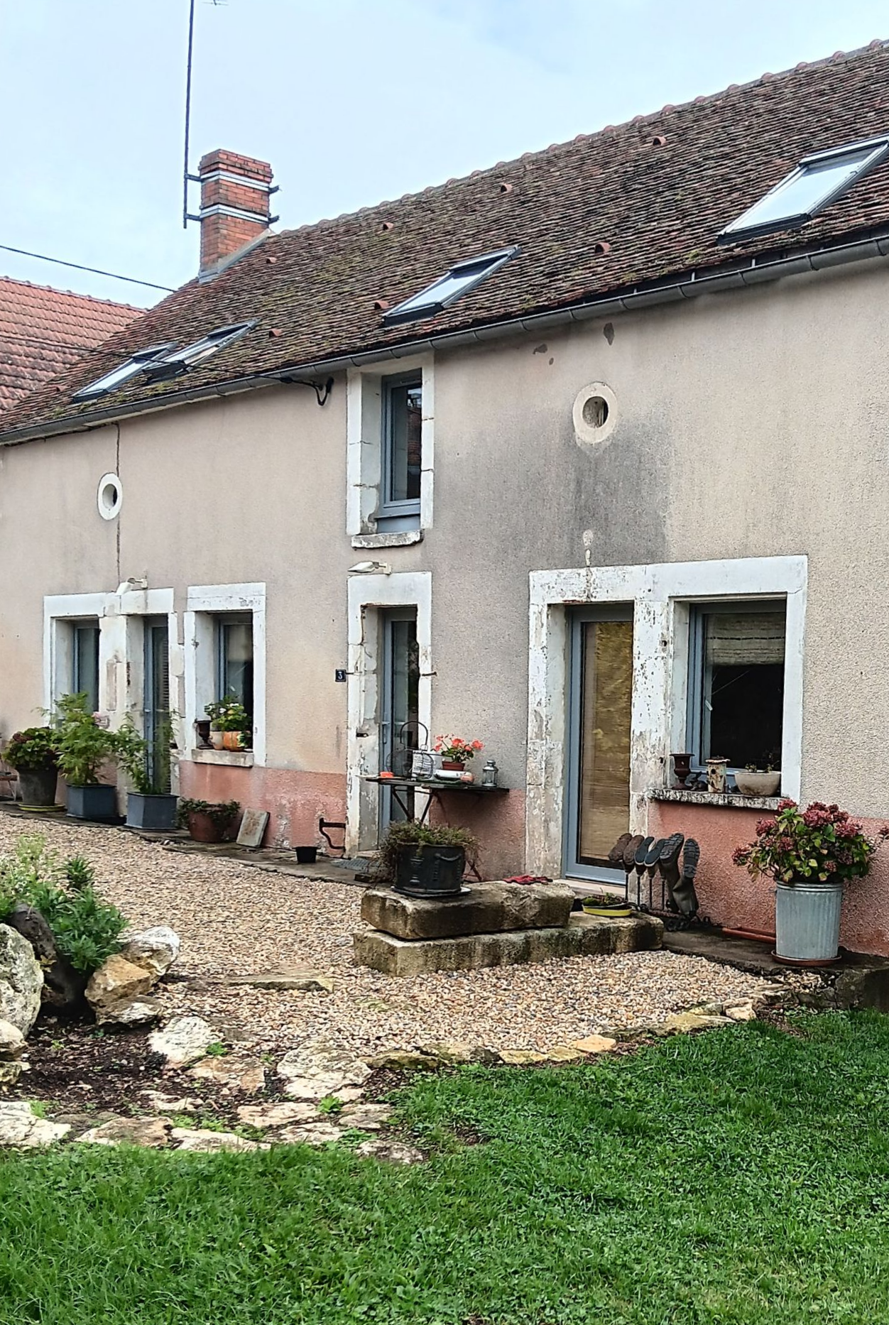 Une maison rustique en pierre beige, à deux étages, avec un toit de tuiles, de nombreuses fenêtres et des pots de fleurs, sur une cour gravillonnée.