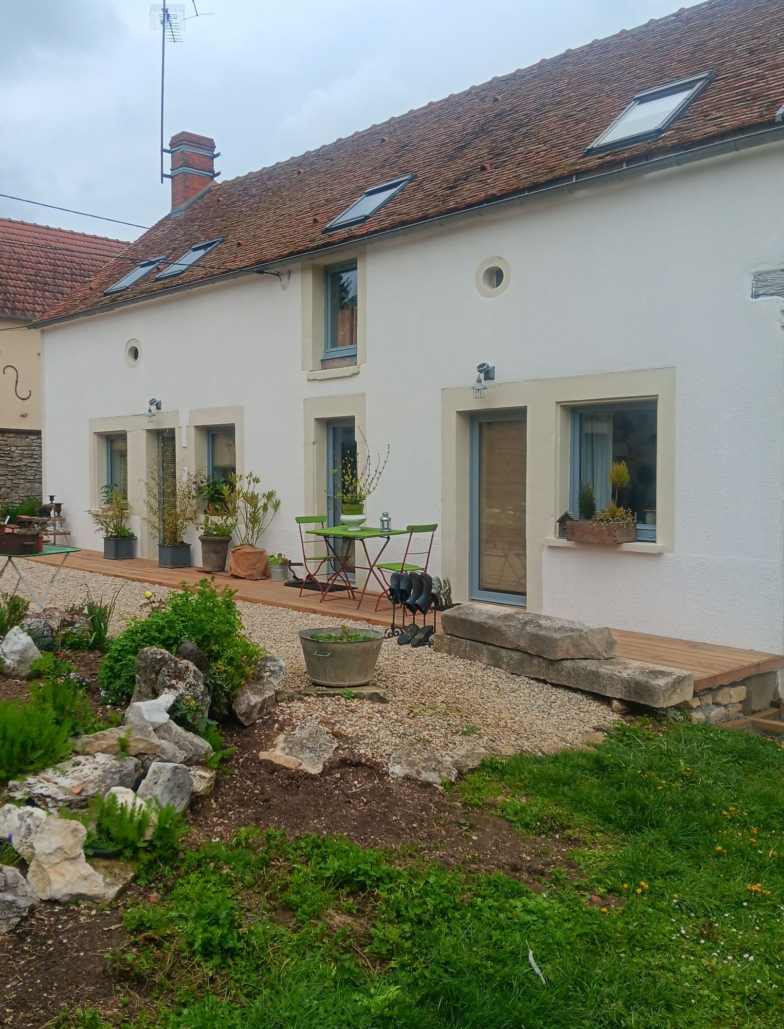 Une maison blanche avec un toit de tuiles, une terrasse pavée et un jardin paysager rocailleux au premier plan.