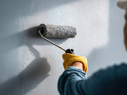 Une personne portant un gant de travail jaune utilise un rouleau à peinture pour appliquer une peinture claire sur un mur intérieur texturé.
