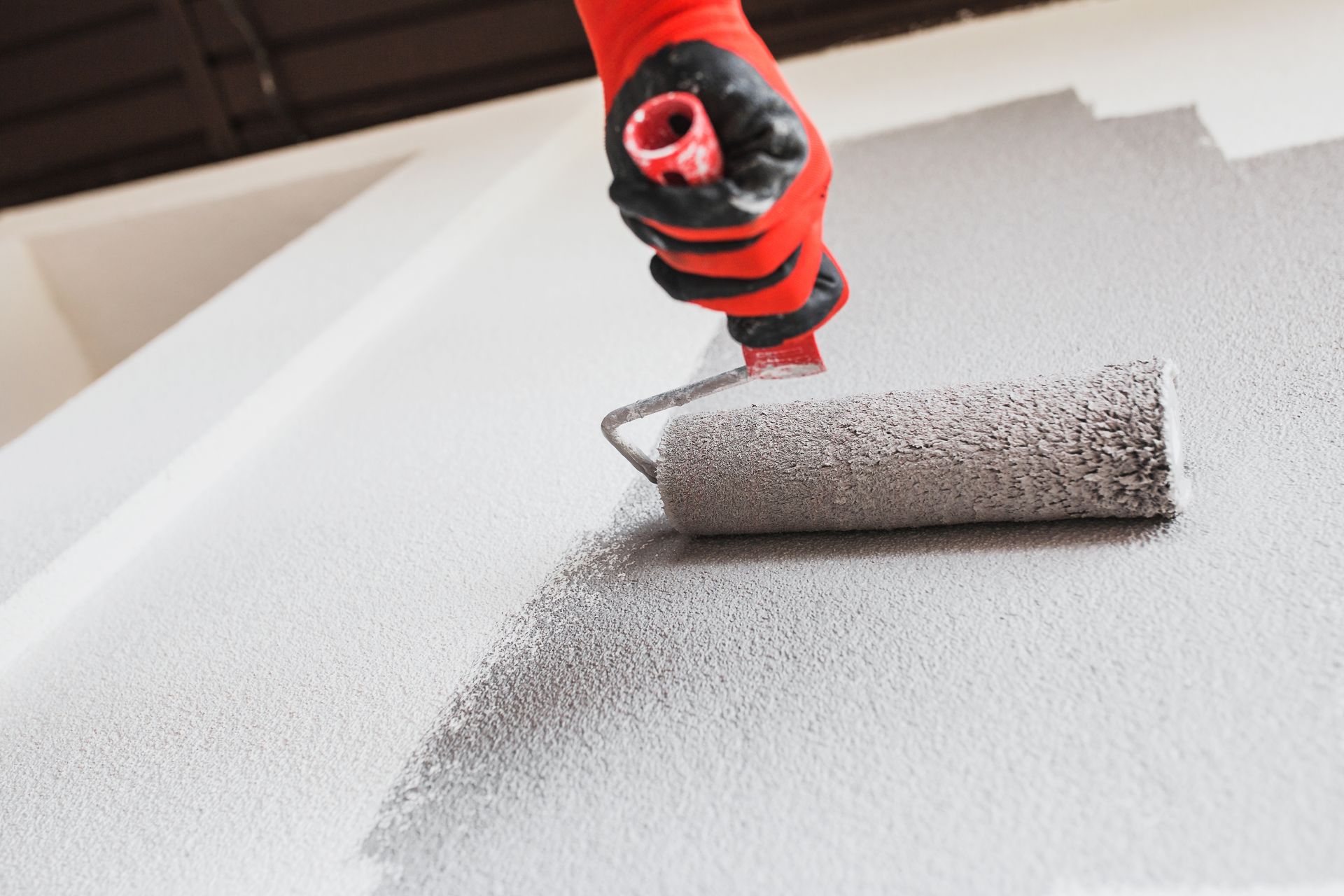 Une personne portant des gants rouges et noirs utilise un rouleau à peinture pour appliquer de la peinture grise sur un mur.