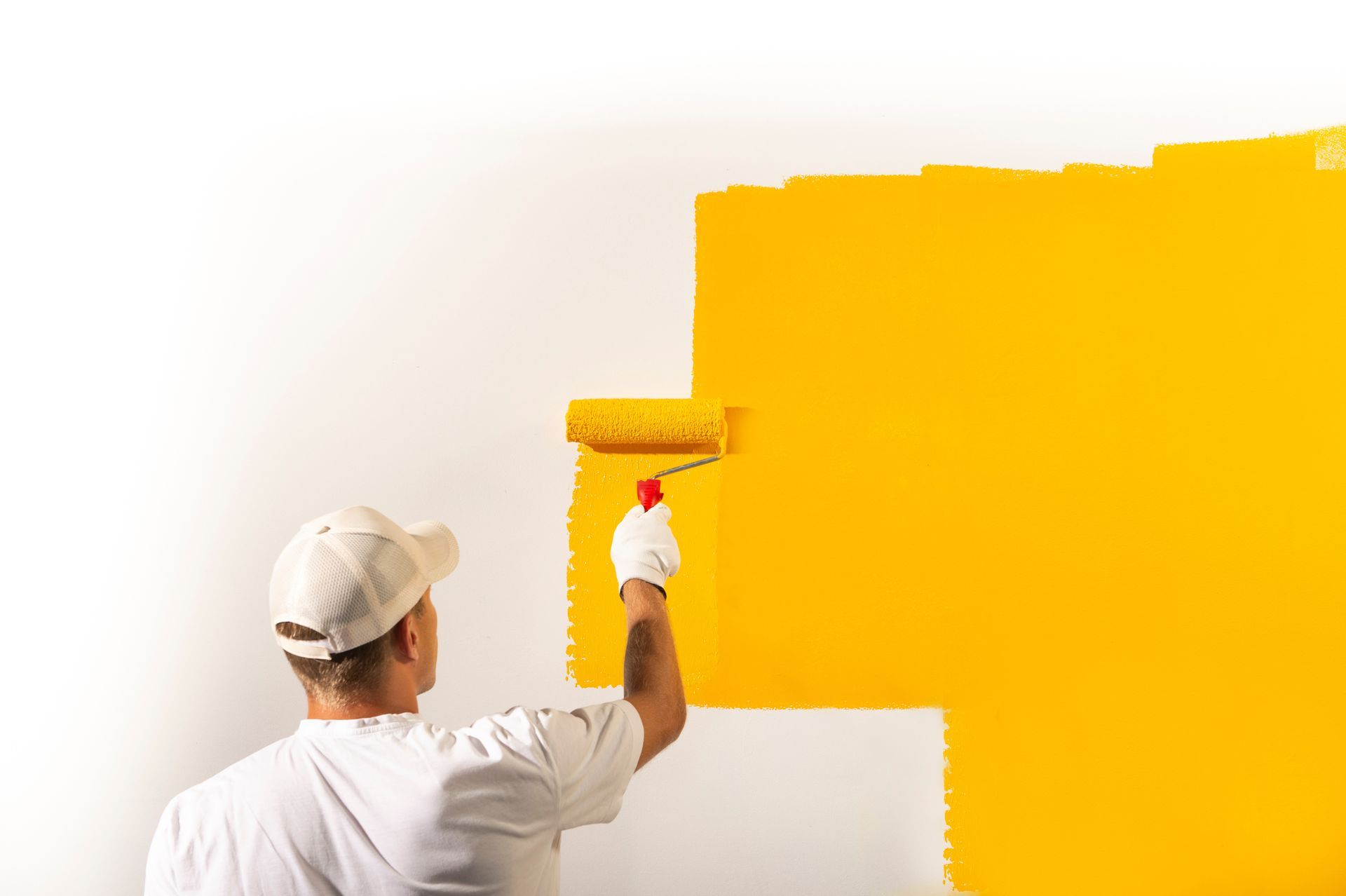 Une personne vêtue d'une chemise et d'une casquette blanches utilise un rouleau à peinture pour appliquer de la peinture jaune vif sur un mur blanc.