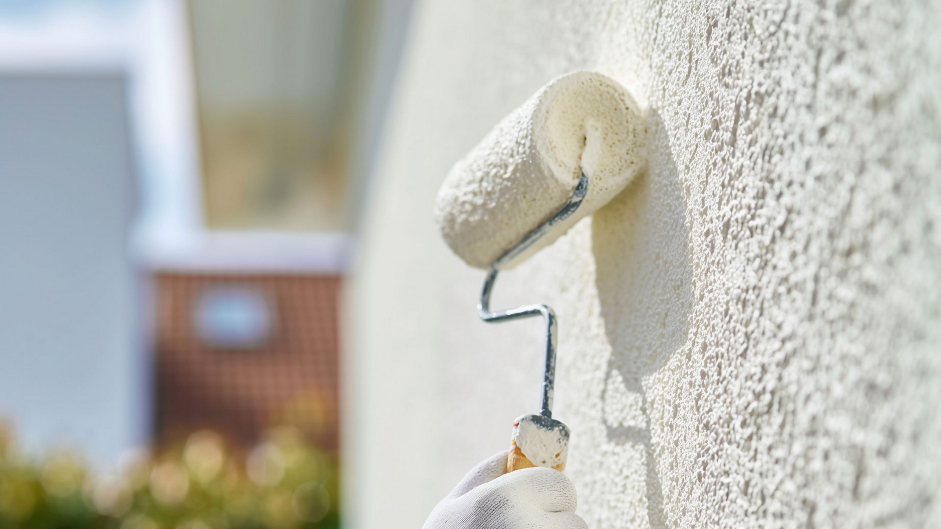 Un peintre en tenue de travail blanche peint un mur extérieur jaune vif à côté d'une porte blanche et d'une petite échelle.