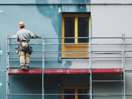 Un ouvrier du bâtiment, casque de chantier sur la tête, se tient sur un échafaudage métallique en train de peindre un mur extérieur bleu près d'une fenêtre jaune.