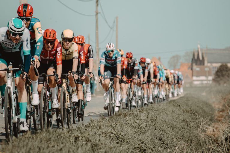 Ciclistas con maillots coloridos pedalean juntos por una carretera soleada. Césped verde y edificios al fondo.