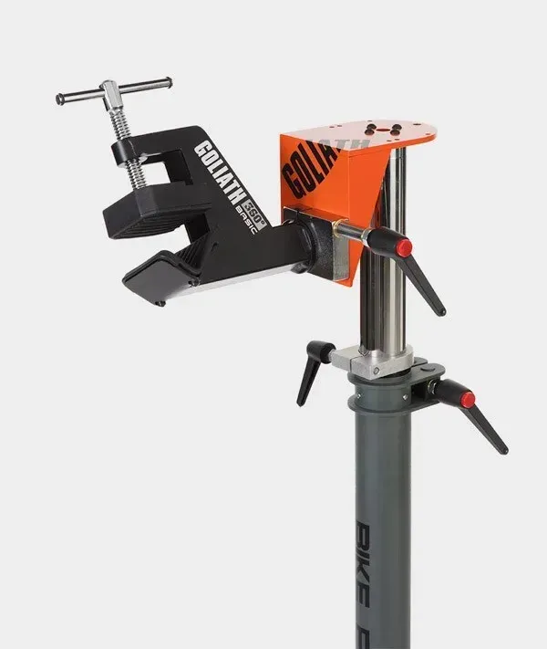 Caballete para reparación de bicicletas con abrazadera negra que sujeta una pieza, logo naranja, poste gris y palancas rojas.