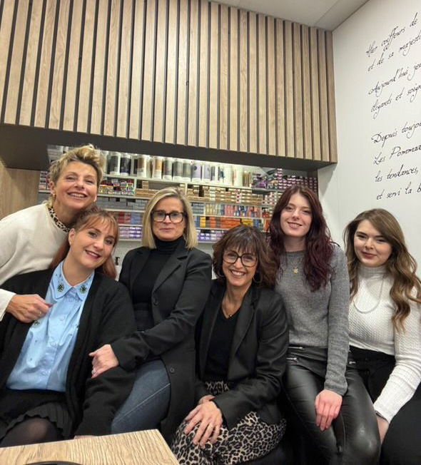Six femmes, assises ou debout, sourient à l'objectif dans un salon de coiffure. Elles sont appuyées contre un mur lambrissé.