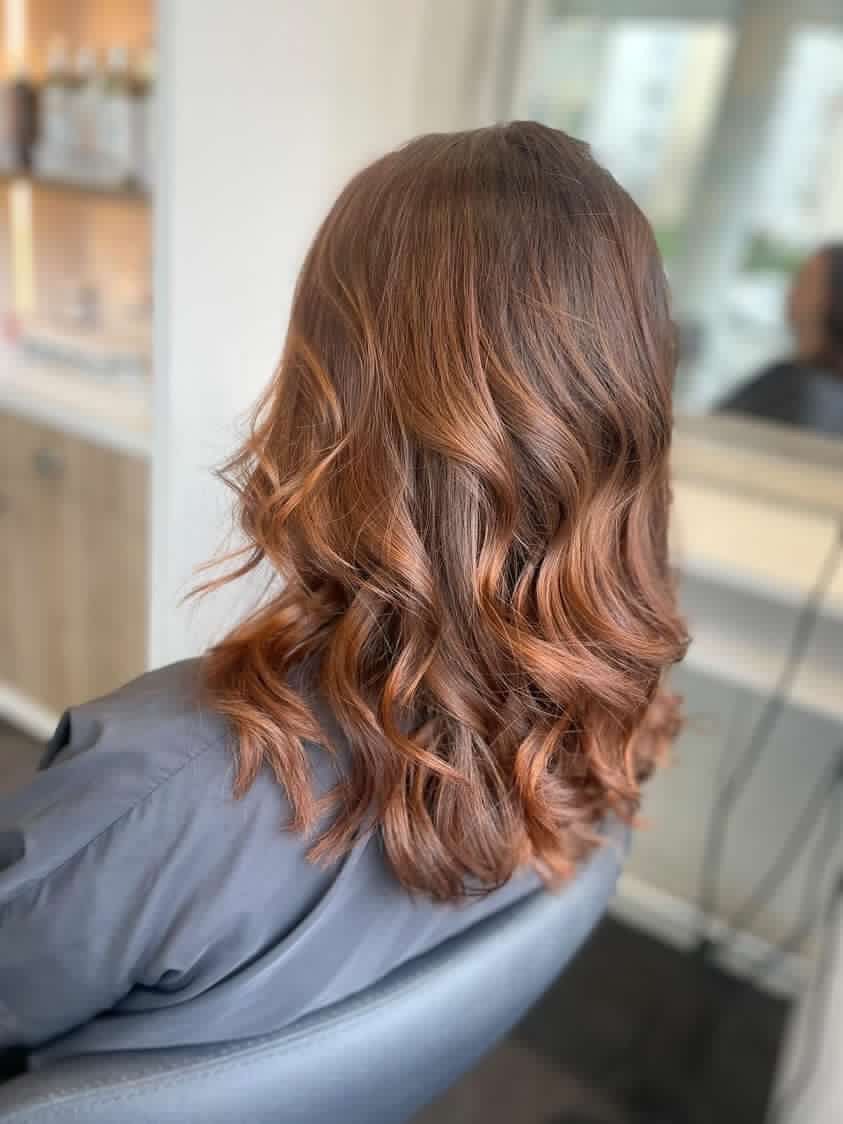 Cheveux bruns avec des ondulations ombrées brun-roux, coiffés en salon, personne assise sur une chaise.