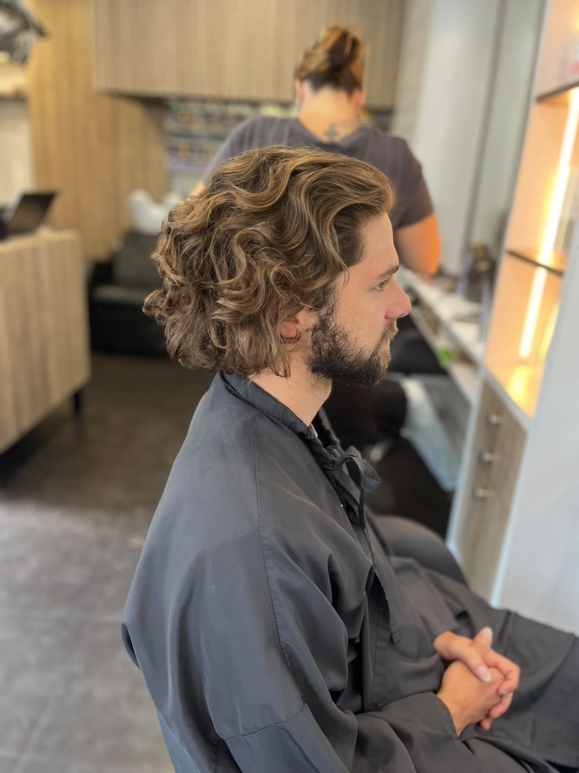 Un homme aux cheveux bouclés, vêtu d'une cape sombre, est assis dans un fauteuil de salon de coiffure. Une autre personne se trouve à l'arrière-plan.