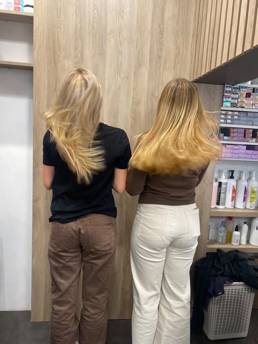 Deux femmes aux coiffures différentes, face à un mur dans un salon de coiffure. L'une porte une chemise noire et un pantalon marron. L'autre un haut marron et un pantalon blanc.