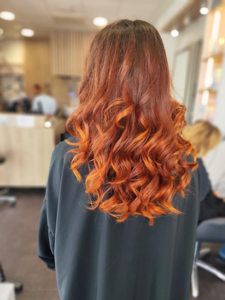 Femme aux longs cheveux ondulés, couleur or rose, à l'extérieur, devant un bâtiment blanc.