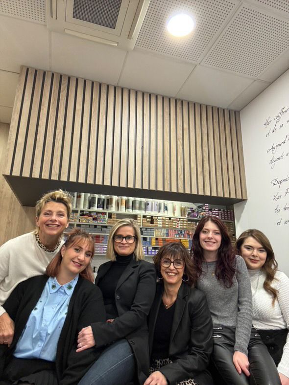 Sept femmes sourient ensemble dans un salon de coiffure. On aperçoit des boiseries claires et des produits sur des étagères.