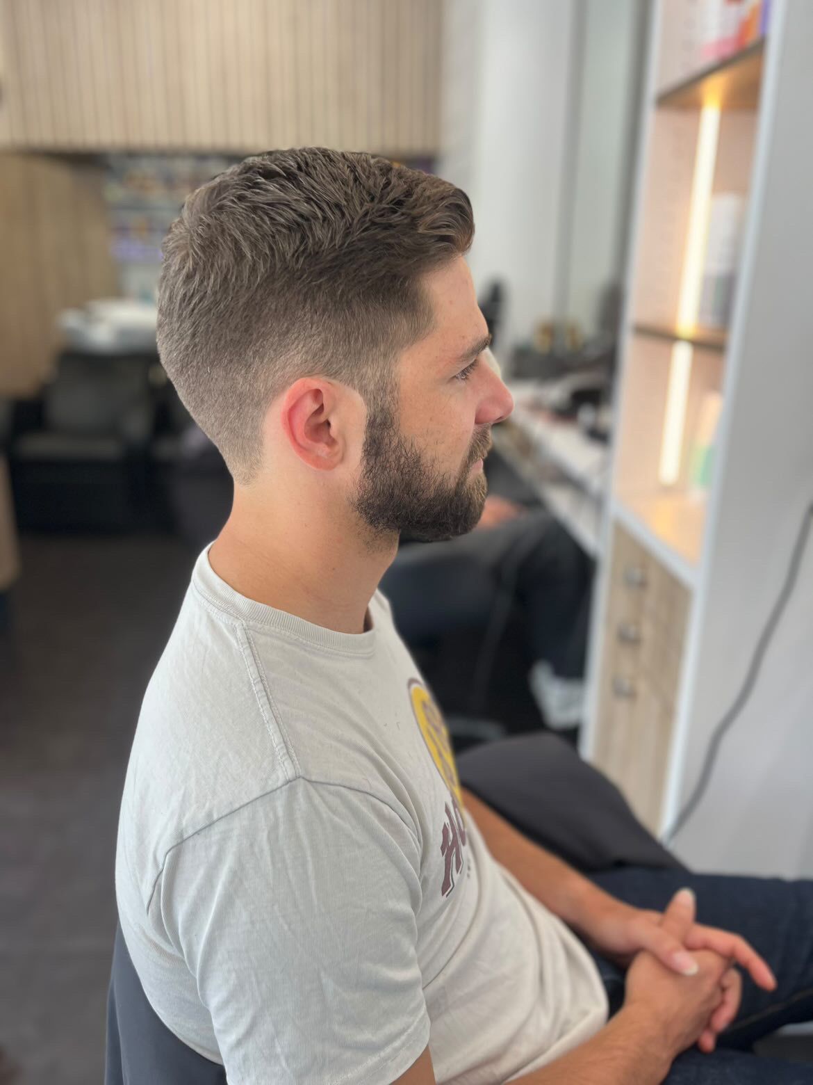Un homme aux cheveux courts et à la barbe fournie se trouve dans un salon de coiffure. Il porte un t-shirt et est assis.