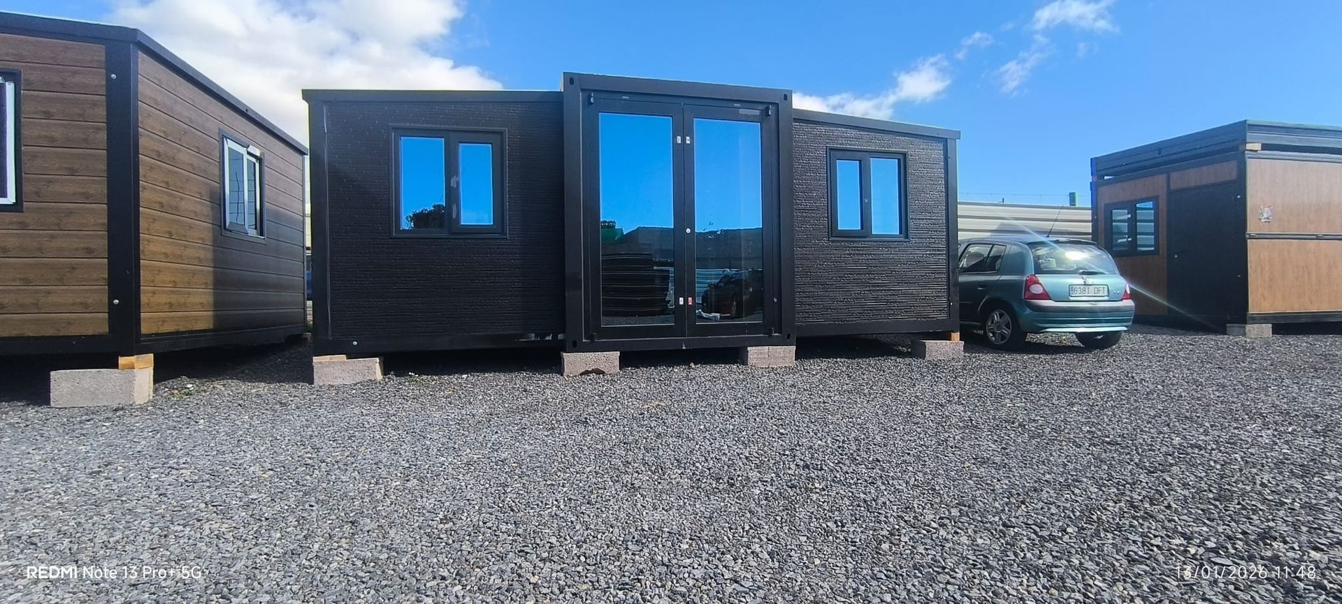 Modernas cabañas modulares negras sobre grava, con un coche aparcado entre dos unidades bajo un cielo despejado.