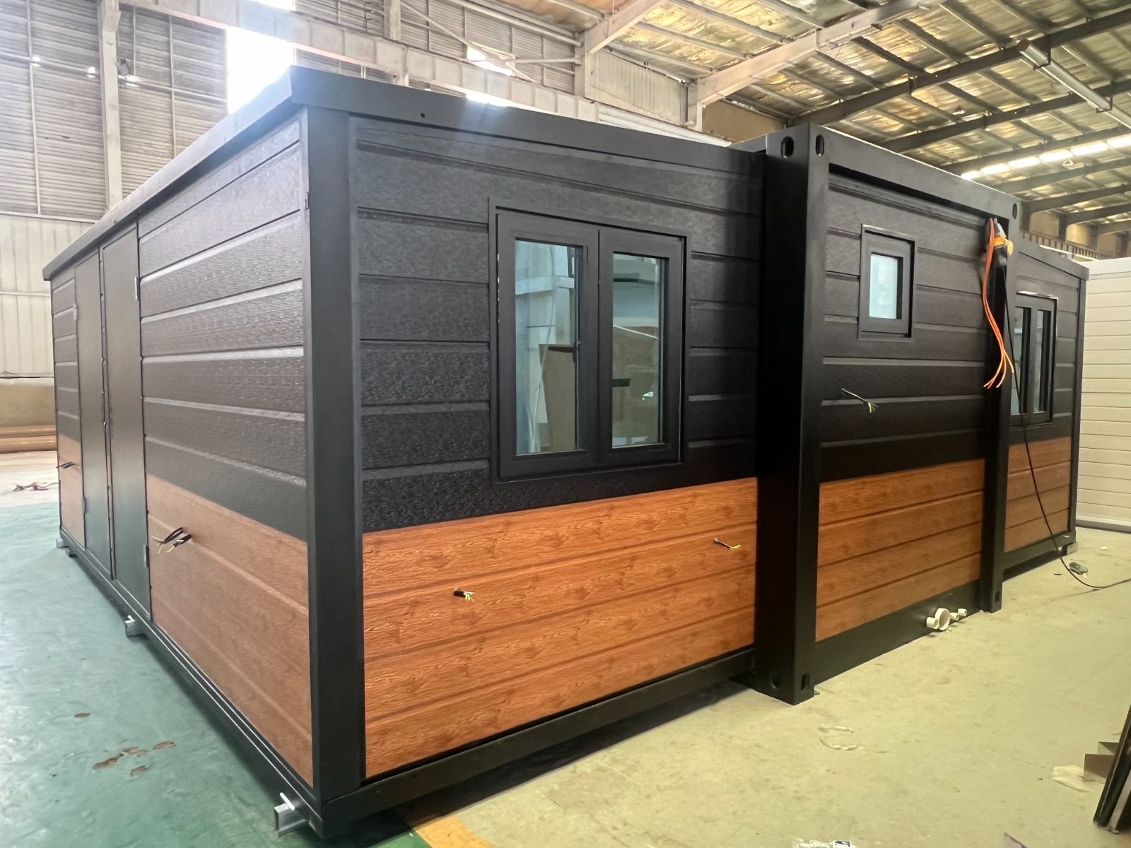 Cabina modular negra con paneles inferiores de madera en un almacén, con una persona de pie a la derecha.