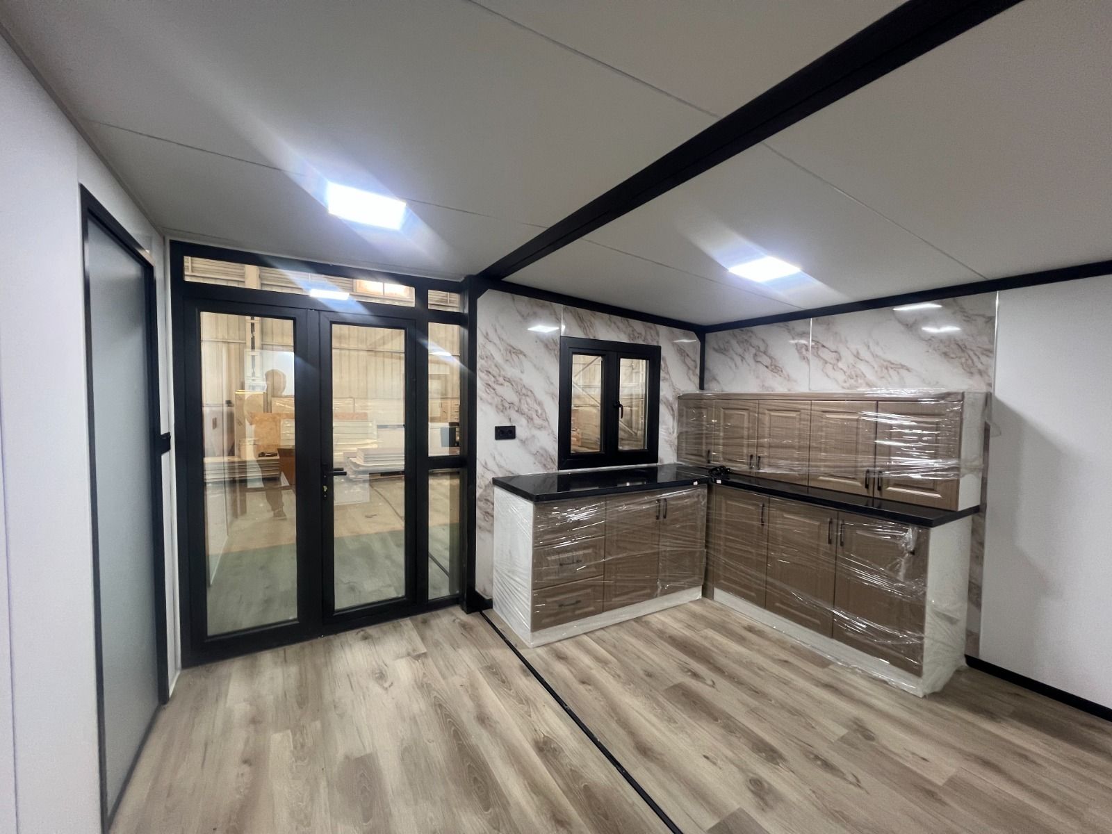 Cocina moderna y vacía con suelo de madera clara, puertas de cristal con marcos negros y encimeras alicatadas.