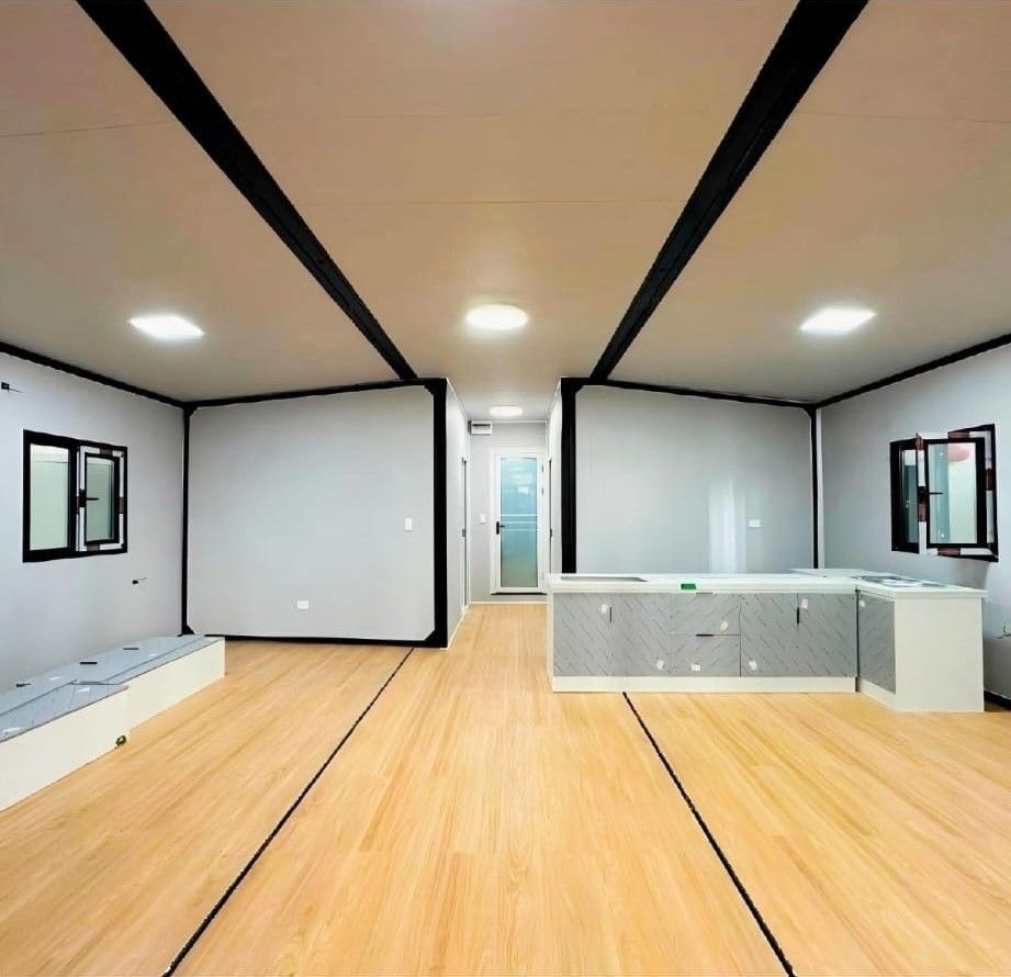 Habitación moderna vacía con suelos de madera clara, paredes blancas, vigas negras en el techo y ventanas a ambos lados.