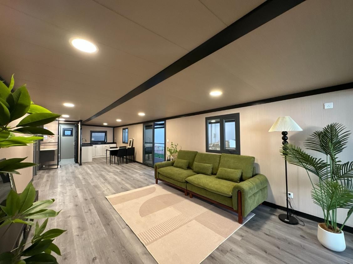 Sala de estar moderna con sofá verde, lámpara de pie, plantas de interior y cocina abierta.