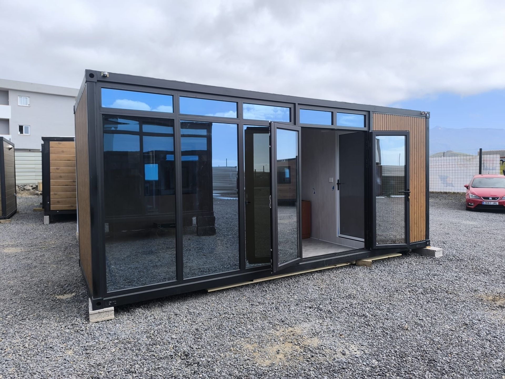 Pequeña construcción modular con estructura negra y grandes ventanales en un terreno de grava al aire libre.