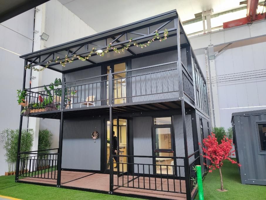 Casa negra de dos plantas con balcones, terraza y plantas en macetas dentro de un espacio tipo almacén.