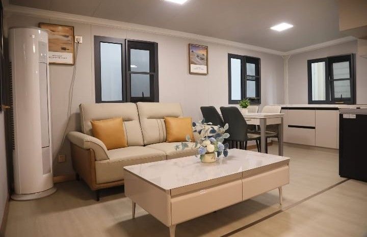 Sala de estar moderna con sofá beige, mesa de centro, ventanas y área de cocina abierta.