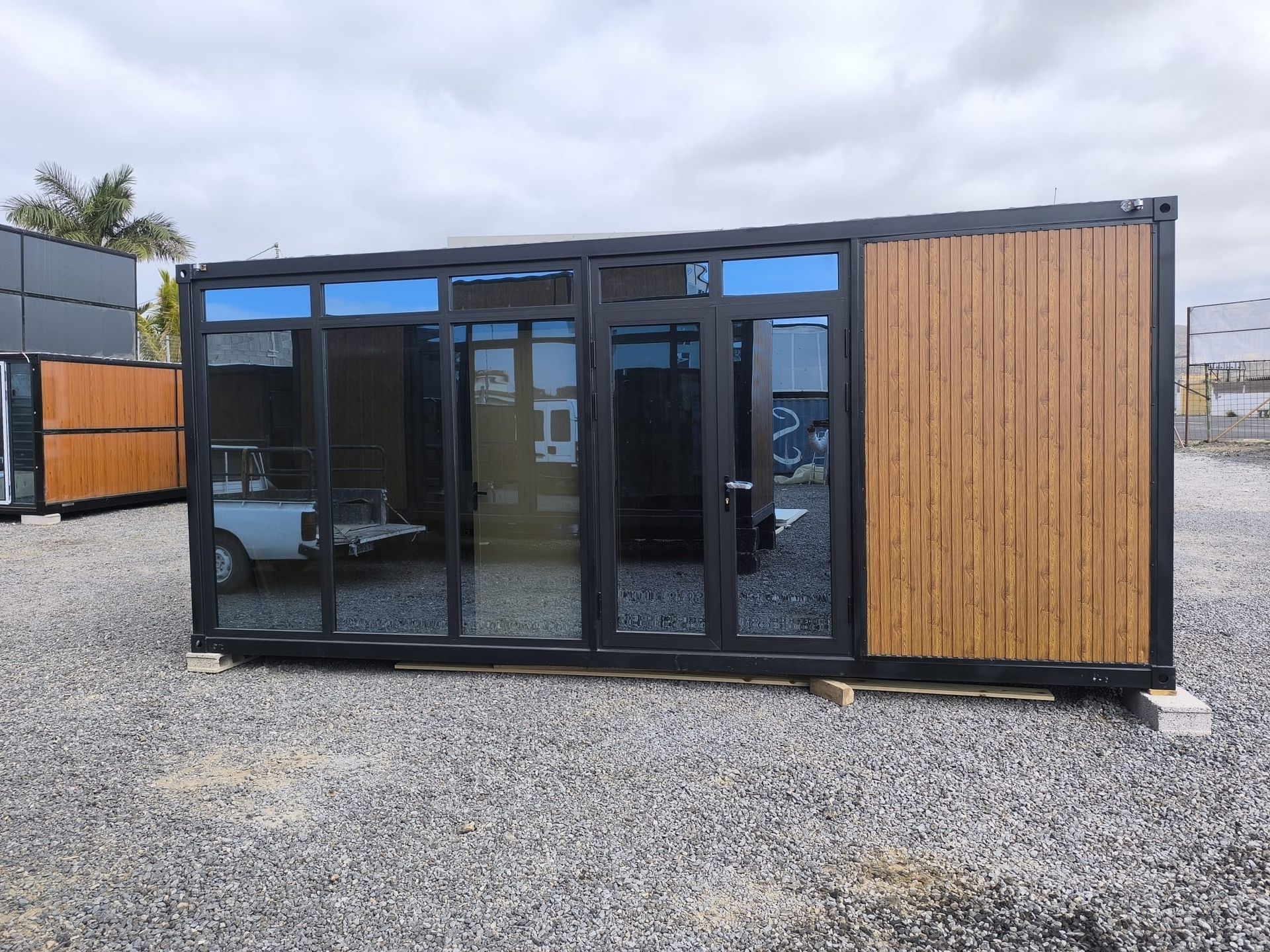 Pequeña cabaña modular moderna de madera y negro con grandes puertas de cristal en un terreno de grava.