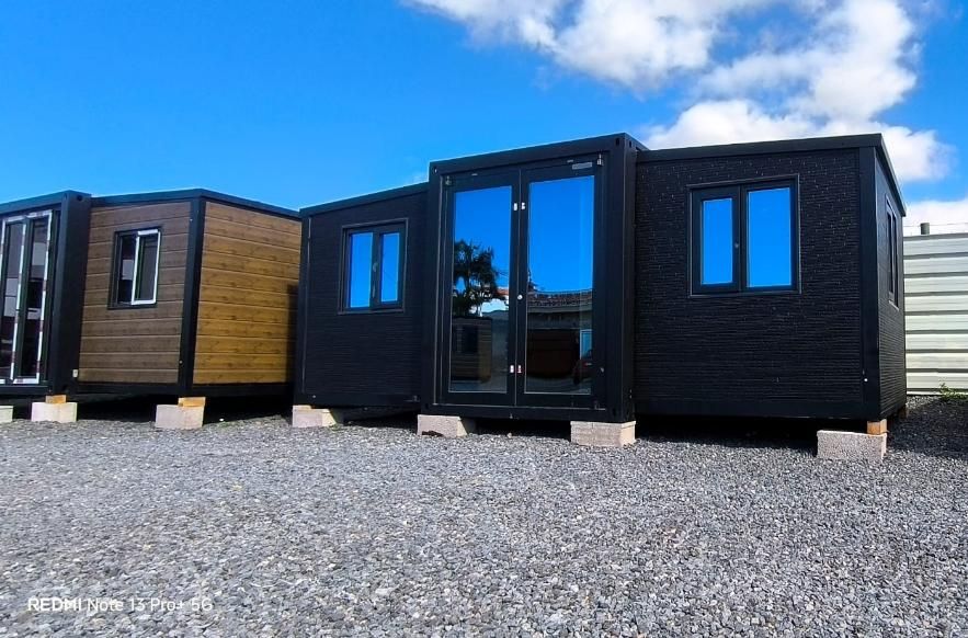 Cabañas modulares negras sobre grava con un coche plateado aparcado y el cielo azul reflejado en las ventanas.