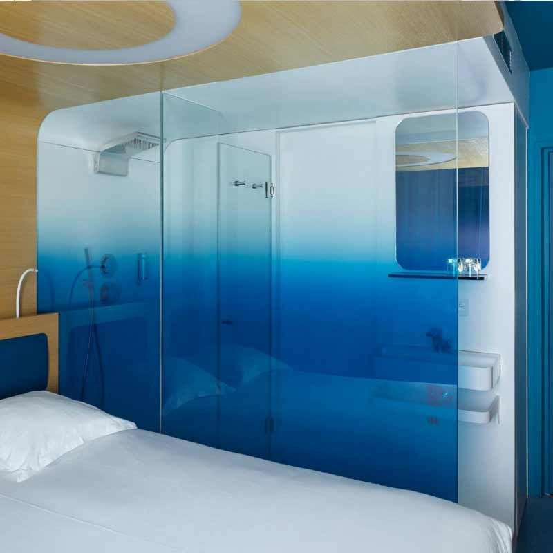 Schlafzimmer mit blau-weiß schimmernder Glasduschkabine. Das Bett ist weiß, die Wand aus Holz.