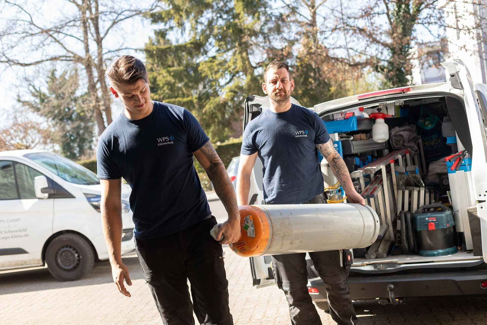 Zwei Männer tragen ein großes Rohr vor einem Lieferwagen.