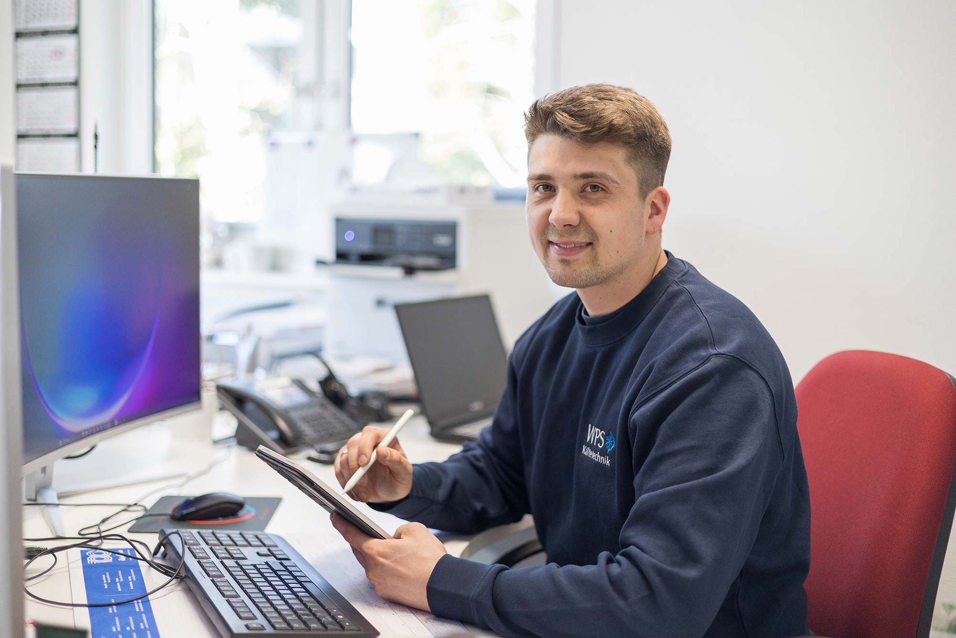 Ein Mann sitzt mit einem Computer und einem Tablet an einem Schreibtisch.