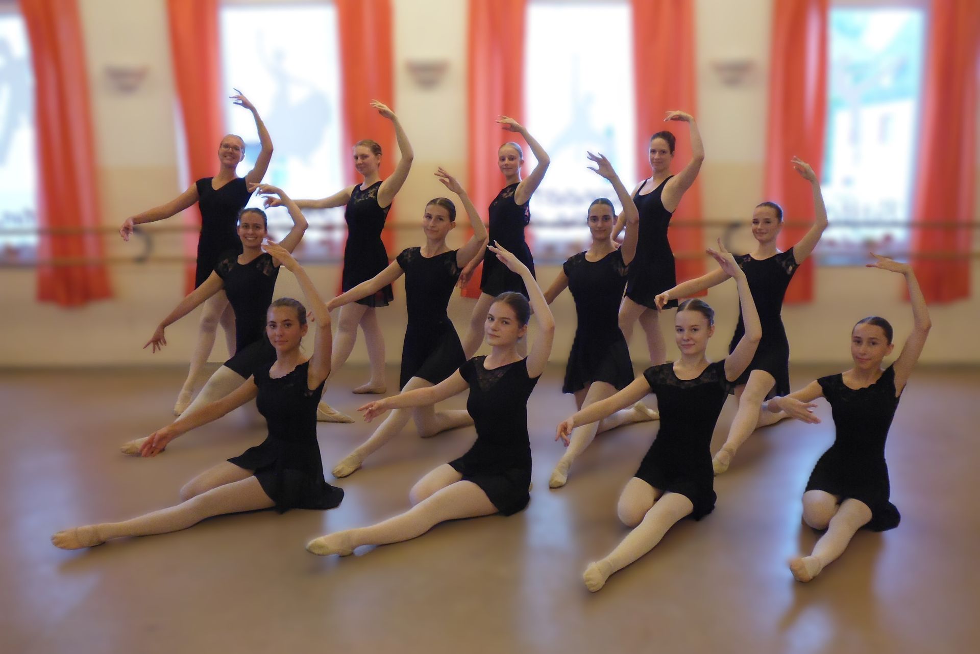 Fünf Tänzerinnen posieren in einem Studio, die Arme in Ballettformation vor einem großen Fenster erhoben.