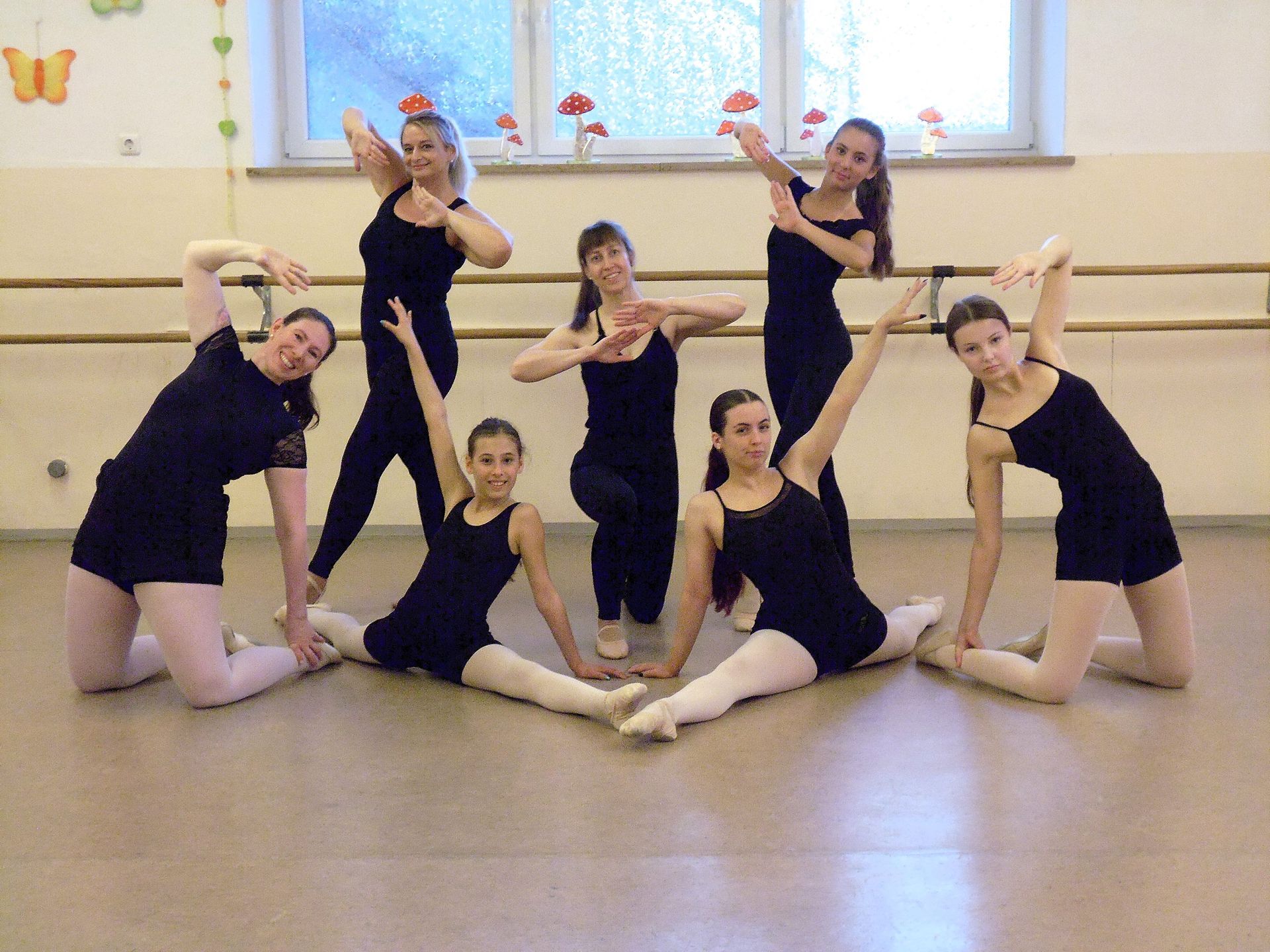 Fünf Tänzerinnen in schwarzen Trikots posieren in einem Studio mit großem Fenster und Ballettstange.