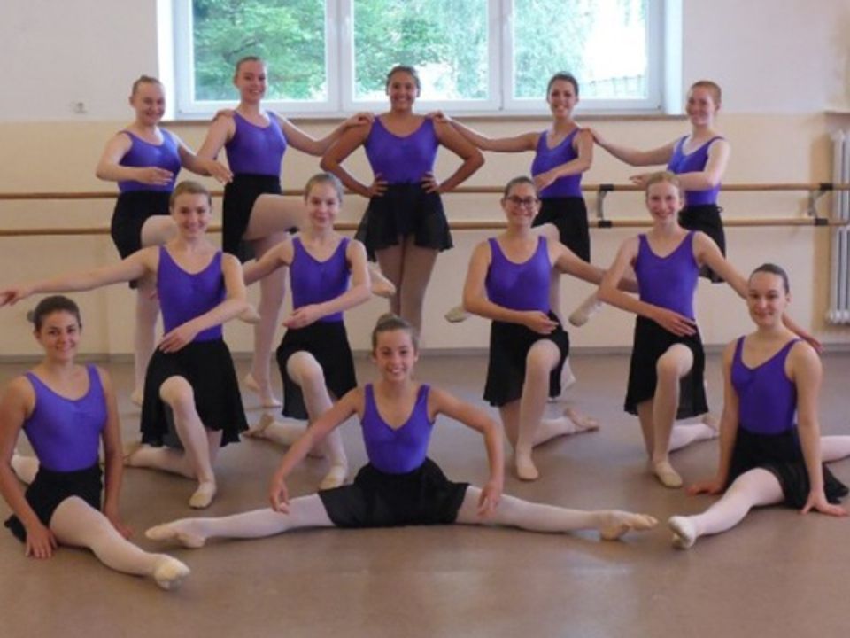 Ballettschülerinnen posieren in einem Tanzstudio, sie tragen lila Oberteile und schwarze Röcke/Trikots.