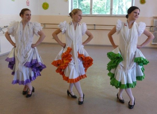 Drei Tänzerinnen in weißen Kleidern mit lila, orangen und grünen Akzenten treten in einem Studio auf.